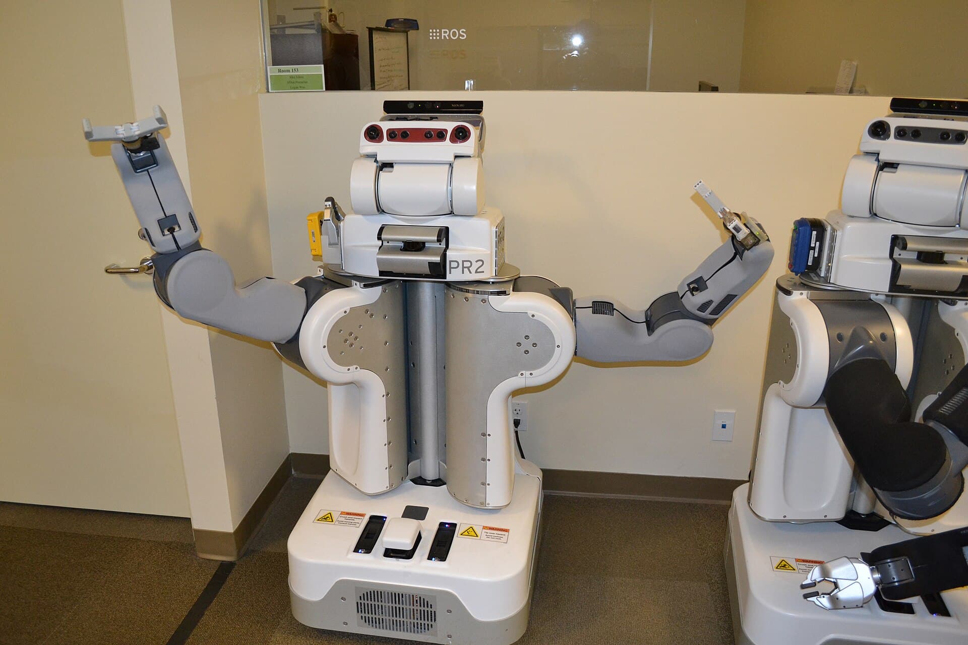 Doi roboți umanoizi de serviciu, albi, expuși într-un spațiu interior; unul este marcat PR2. (Foto de arhivă: Oleg Alexandrov / CC BY-SA 3.0, Wikimedia Commons)