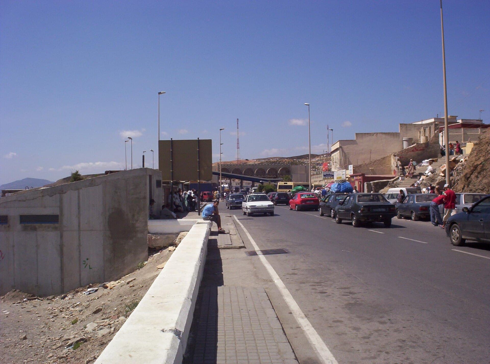 O stradă urbană aglomerată din Ceuta, cu mașini, pietoni și clădiri; în fundal se vede zona de frontieră. (Foto: The original uploader was Jguk 2 at English Wikipedia . / CC BY-SA 3.0, Wikimedia Commons)