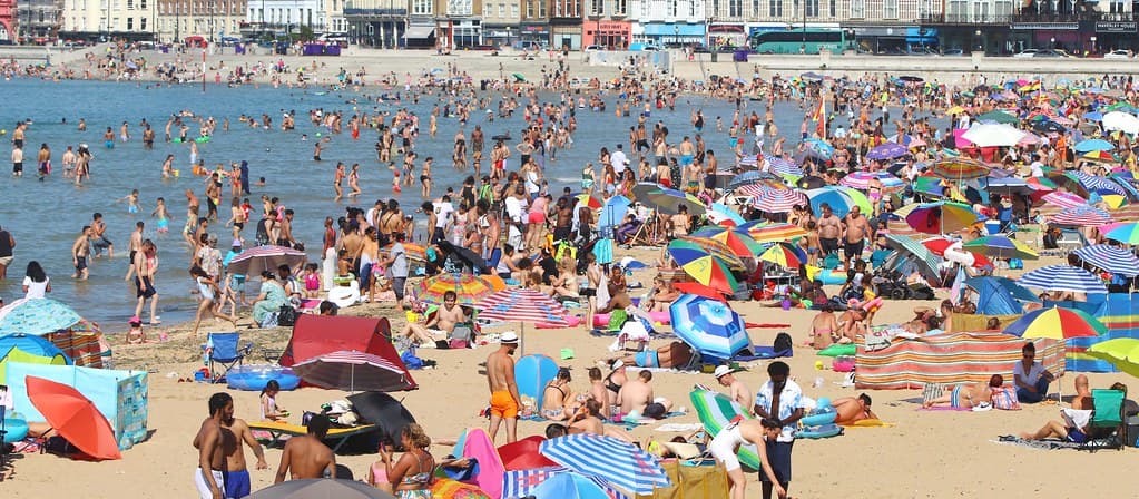 O plajă aglomerată, cu numeroși oameni la soare și în apă, într-o zi călduroasă. (Foto de arhivă: Graham Ó Síodhacháin / BY-SA 2.0, Openverse)