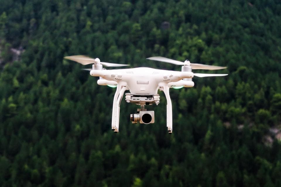O dronă quadcopter albă, cu cameră, care zboară deasupra unei păduri. (Foto ilustrativă: Jason Blackeye / CC0 1.0, Openverse)