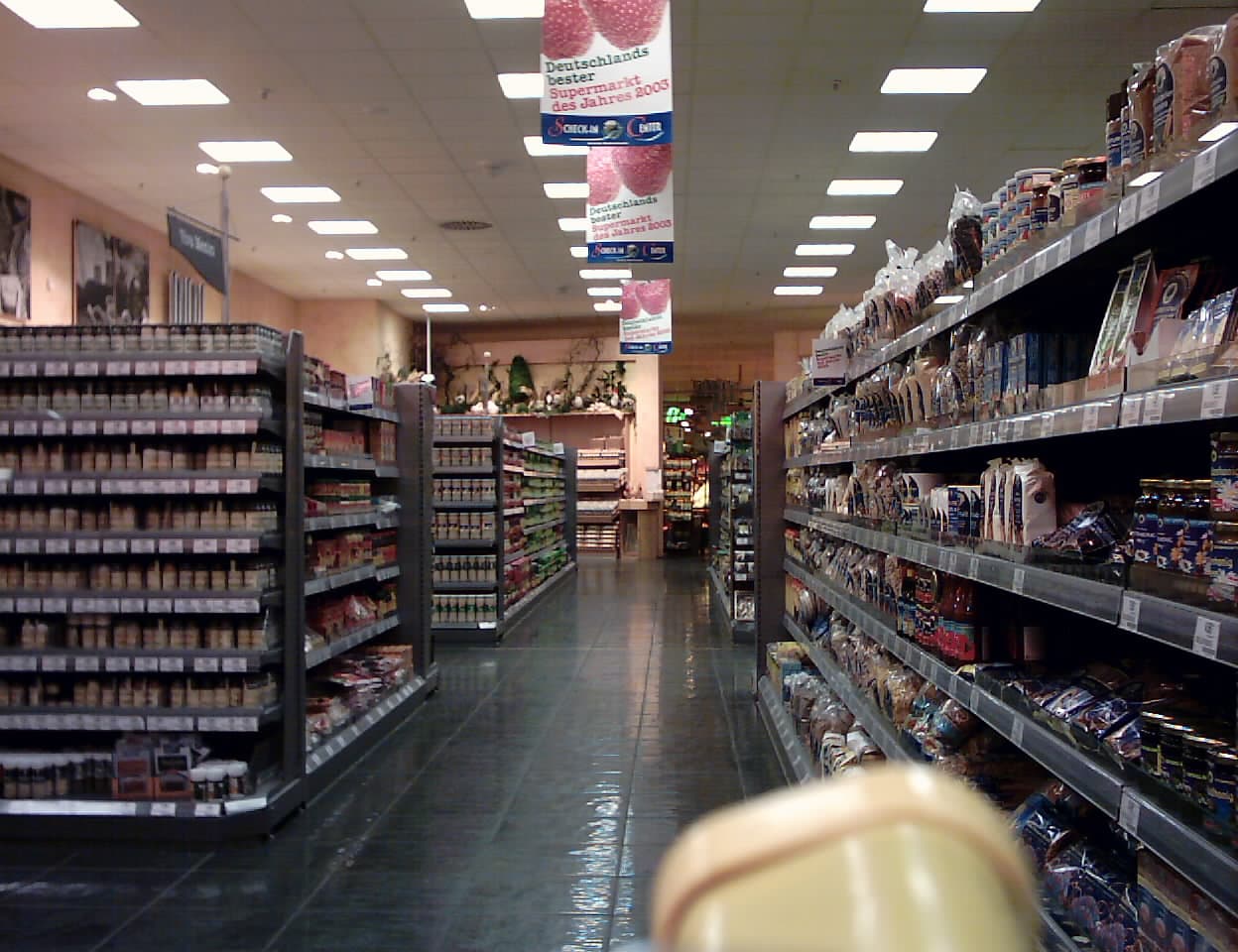 Interiorul unui supermarket, cu rafturi pline de produse și culoar central. (Foto de arhivă: Rob / CC BY-SA 3.0, Wikimedia Commons)