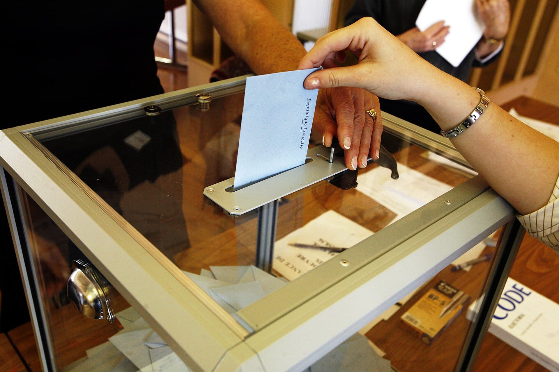 O persoană introduce un buletin de vot într-o urnă transparentă, într-un context electoral. (Foto de arhivă: Rama / CC BY-SA 2.0 fr, Wikimedia Commons)