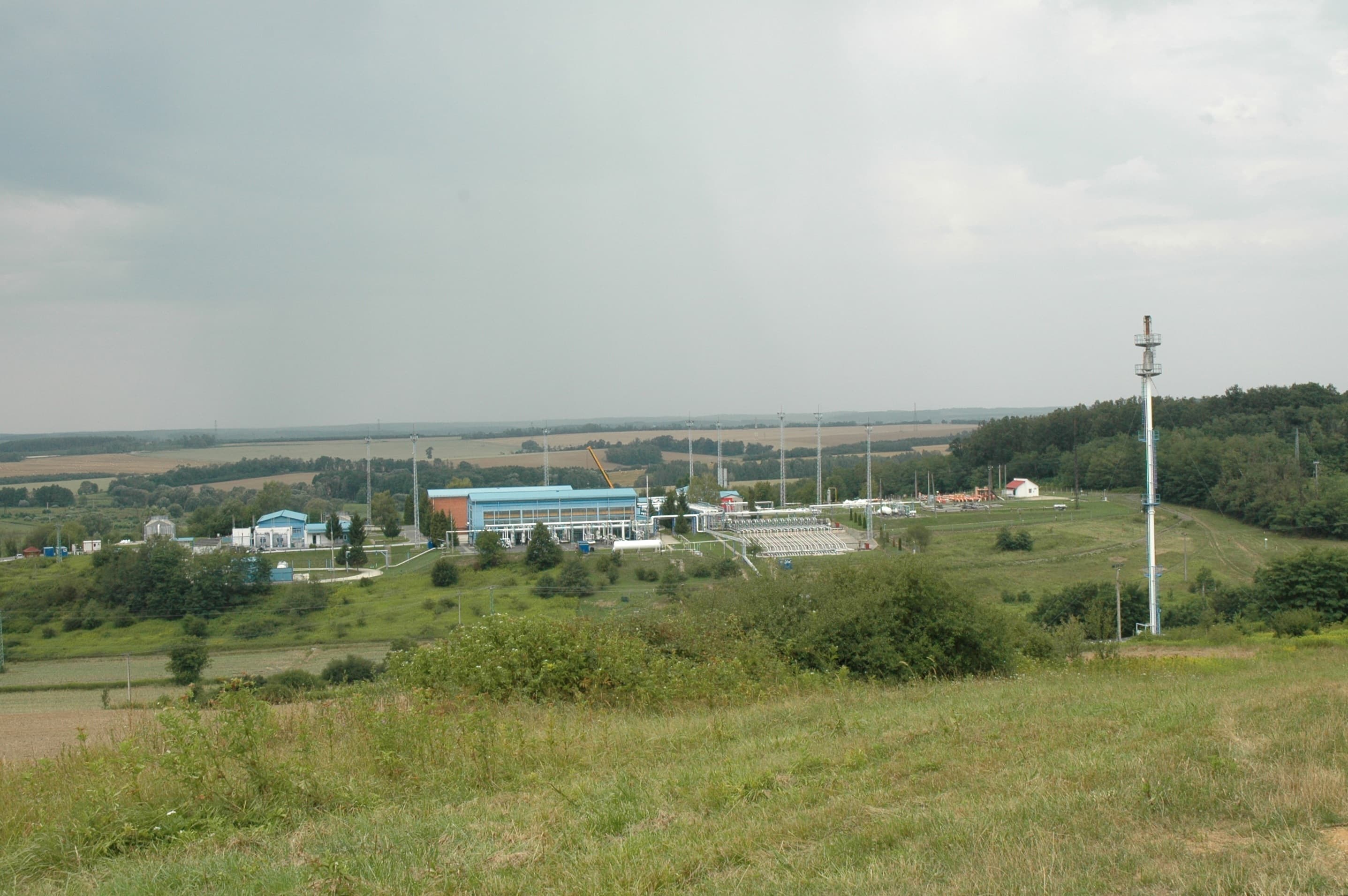 Peisaj rural cu o instalație industrială și conducte sau linii de transport, văzute de la distanță. (Foto de arhivă: VargaA / BY-SA 3.0, Openverse)