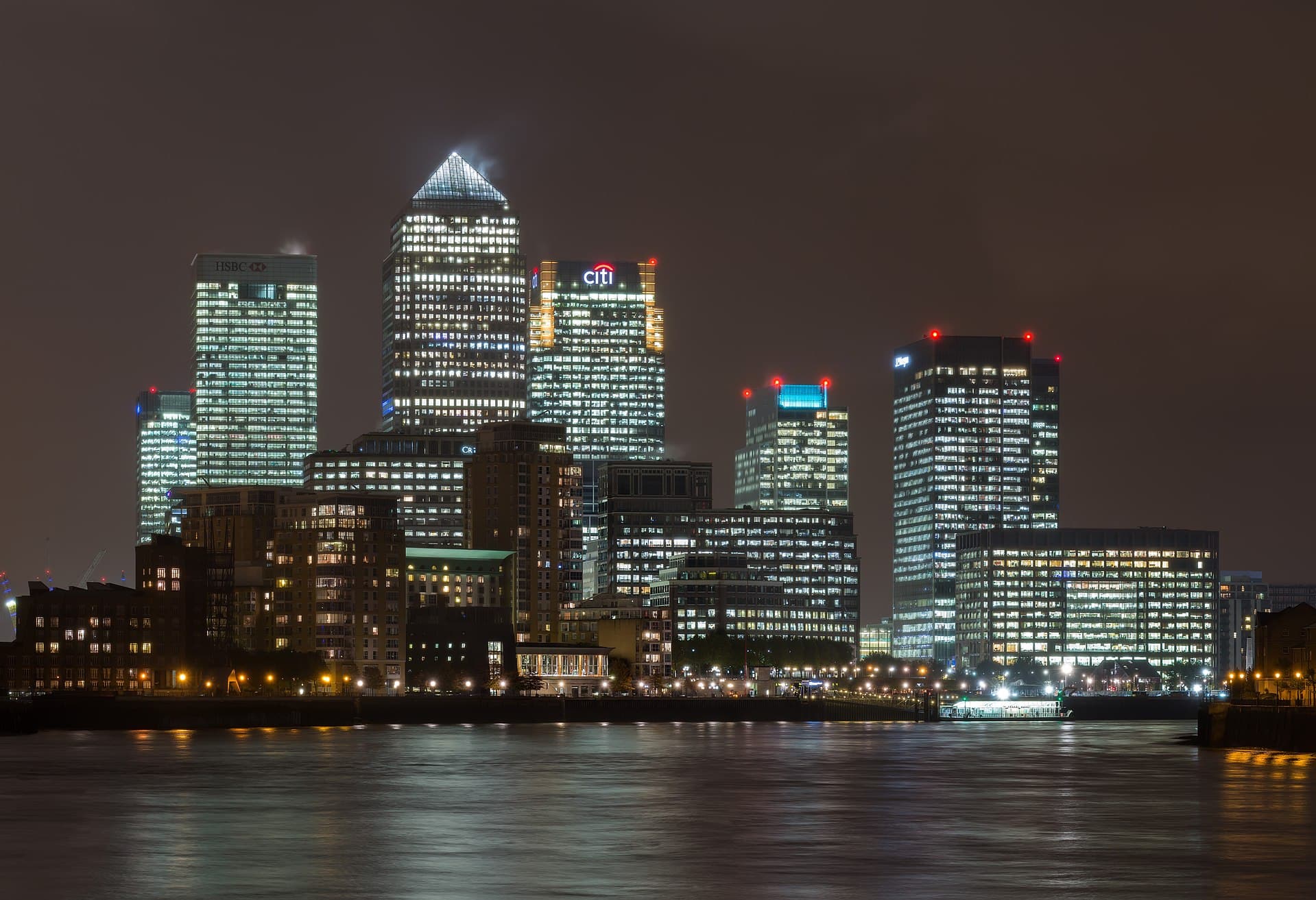 Panorama nocturnă a districtului financiar Canary Wharf din Londra, cu clădiri de birouri iluminate și râul Tamisa în prim-plan. (Foto: Diliff / CC BY-SA 3.0, Wikimedia Commons)