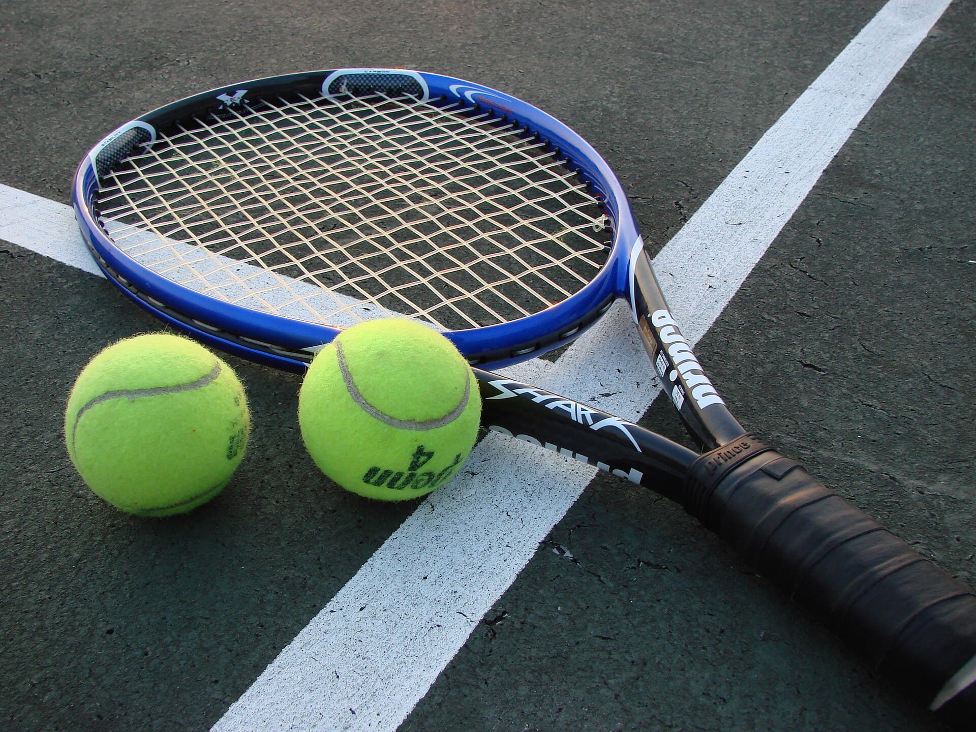 O rachetă de tenis și două mingi de tenis pe un teren de tenis. (Foto de arhivă: Vladsinger at English Wikipedia / CC BY-SA 3.0, Wikimedia Commons)