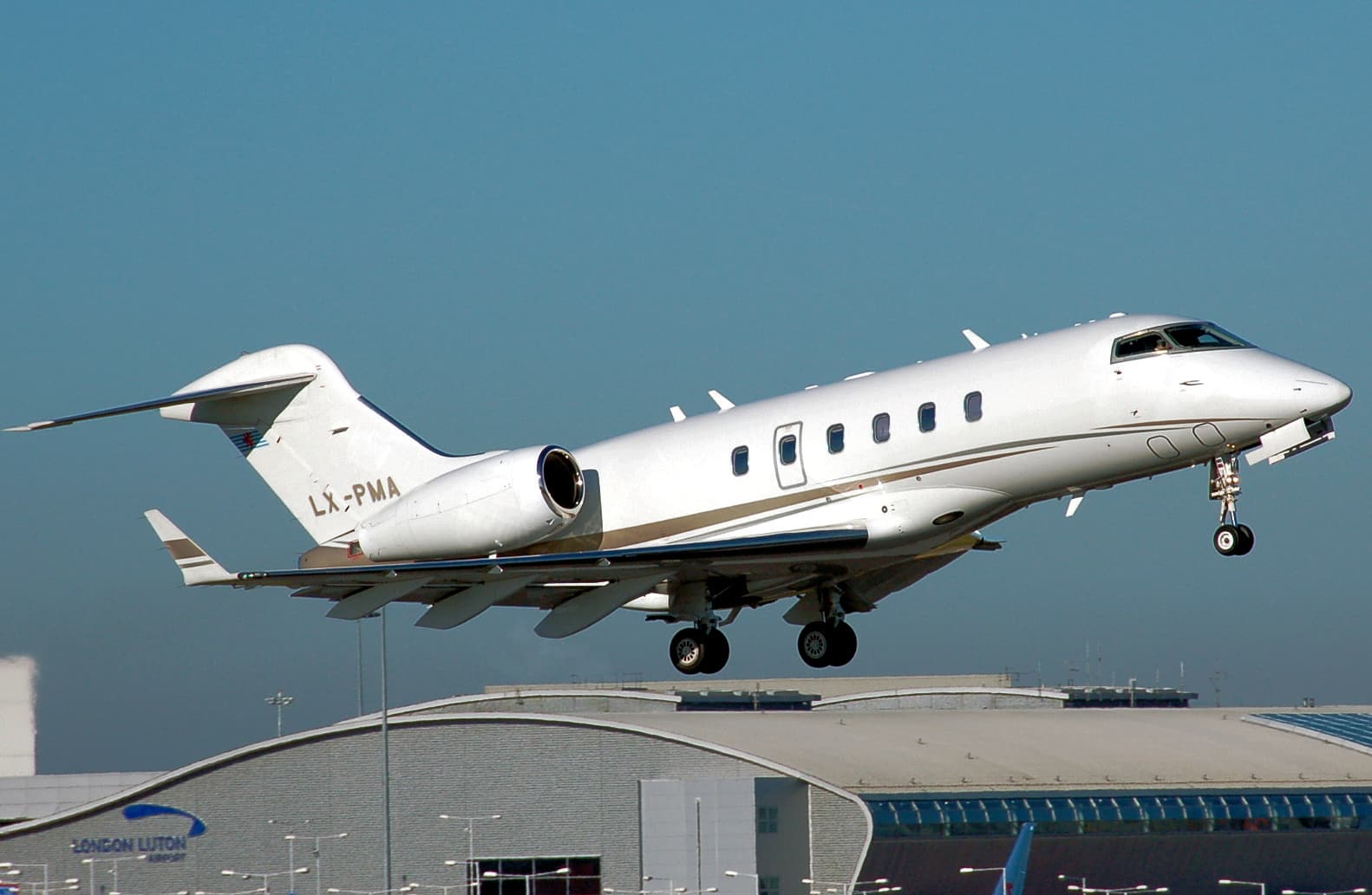 Un avion de afaceri Bombardier alb, în zbor deasupra unei clădiri aeroportuare. (Foto: Arpingstone / Public domain, Wikimedia Commons)