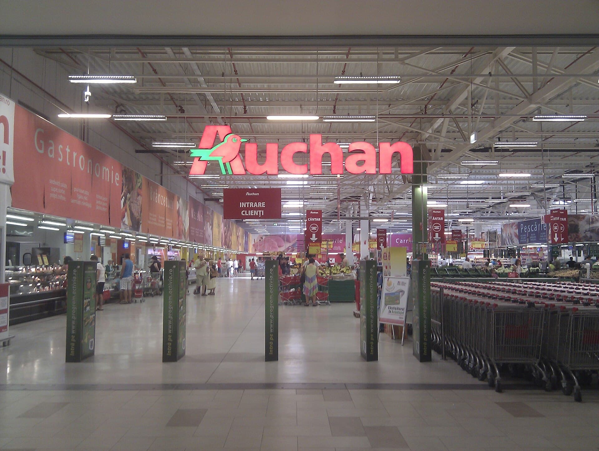 Interiorul unui hipermarket Auchan, cu sigla magazinului, raioane și cărucioare de cumpărături. (Foto: Fieldafar / CC BY 3.0, Wikimedia Commons)