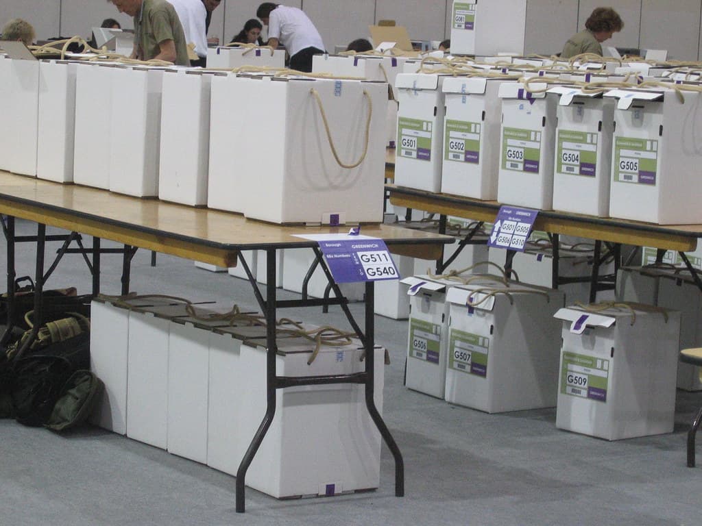 O sală cu urne sau cutii electorale etichetate și persoane care pregătesc materiale pentru alegeri. (Foto de arhivă: secretlondon123 / BY-SA 2.0, Openverse)