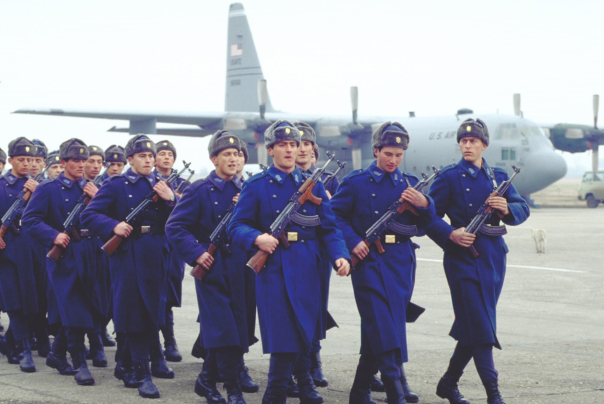 Militari în uniforme albastre, defilând pe un aeroport, cu un avion militar în fundal. (Foto: necunoscut / Public domain, Wikimedia Commons)