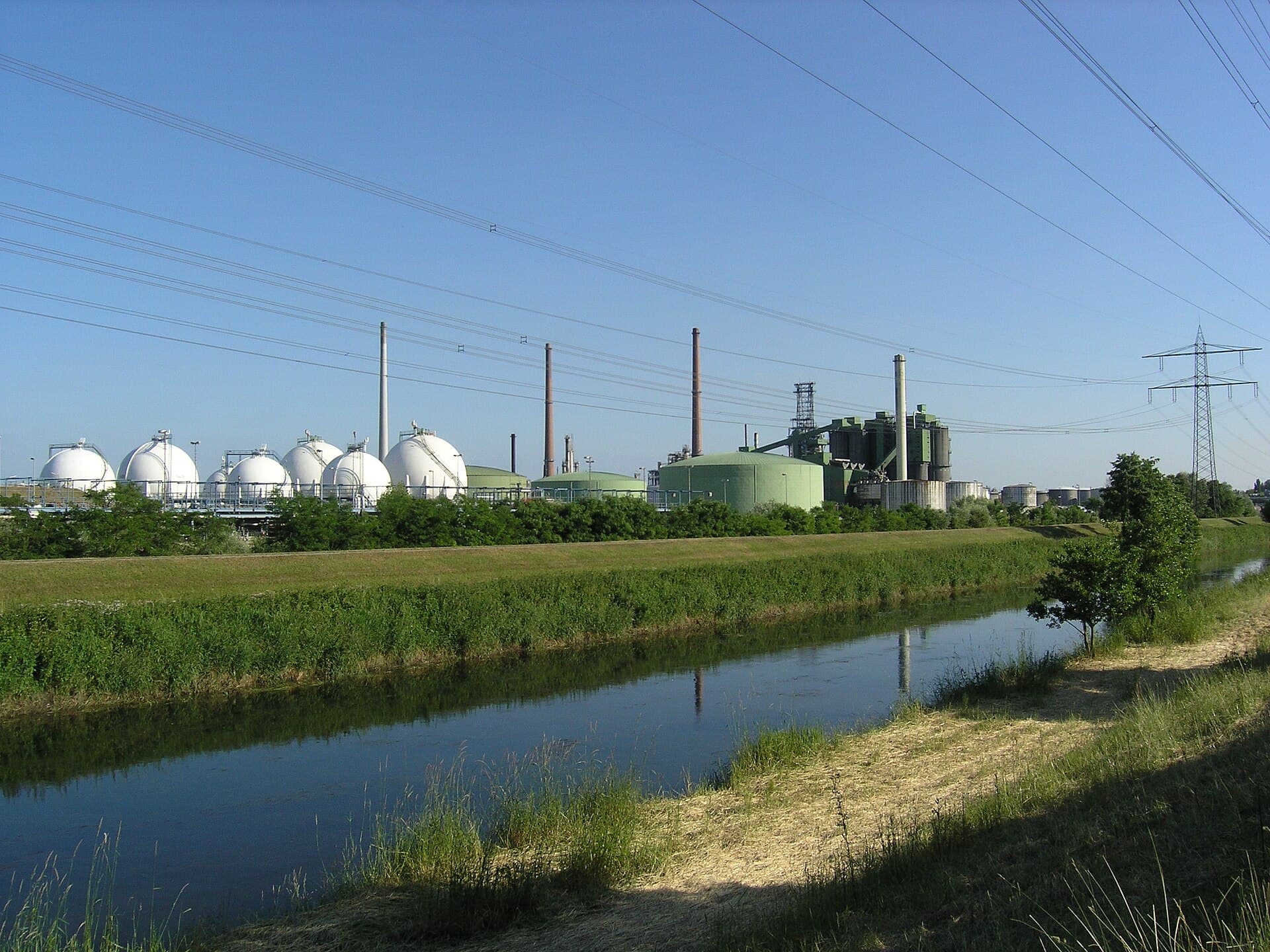 Se vede o instalație industrială energetică, cu rezervoare cilindrice, conducte și linii electrice, fotografiată de lângă un canal. (Foto: Ikar.us ( talk ) / CC BY 2.0 de, Wikimedia Commons)