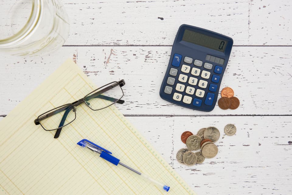 Un calculator, monede, ochelari și un pix pe un caiet, într-un context financiar. (Foto ilustrativă: Artsy Crafty / CC0 1.0, Openverse)