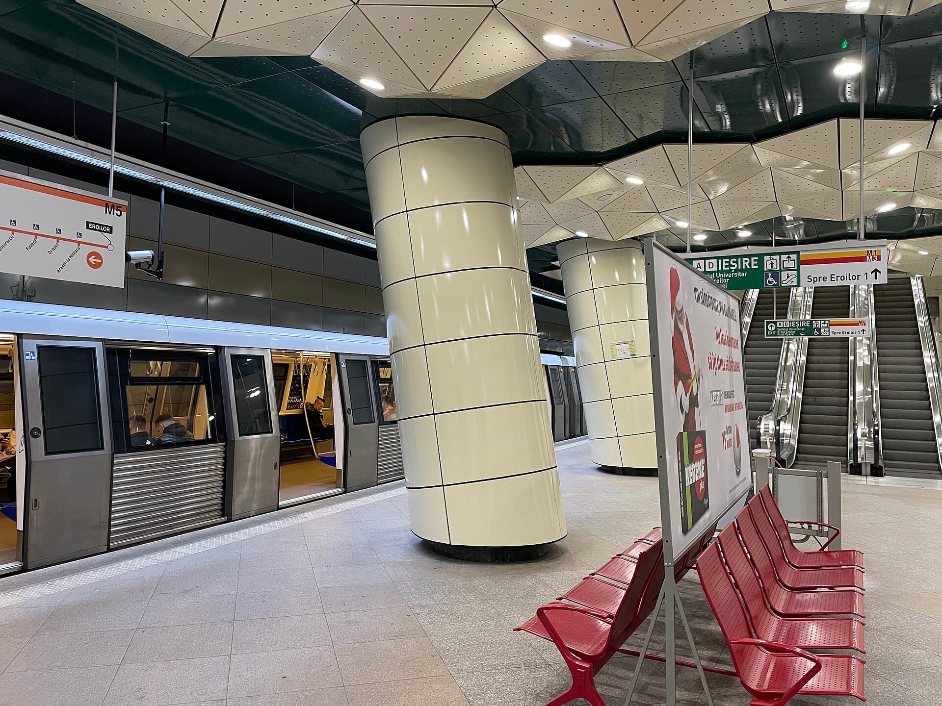 O stație de metrou Metrorex, cu un tren oprit la peron, coloane, scări rulante și indicatoare. (Foto: Puly4q / CC BY-SA 4.0, Wikimedia Commons)
