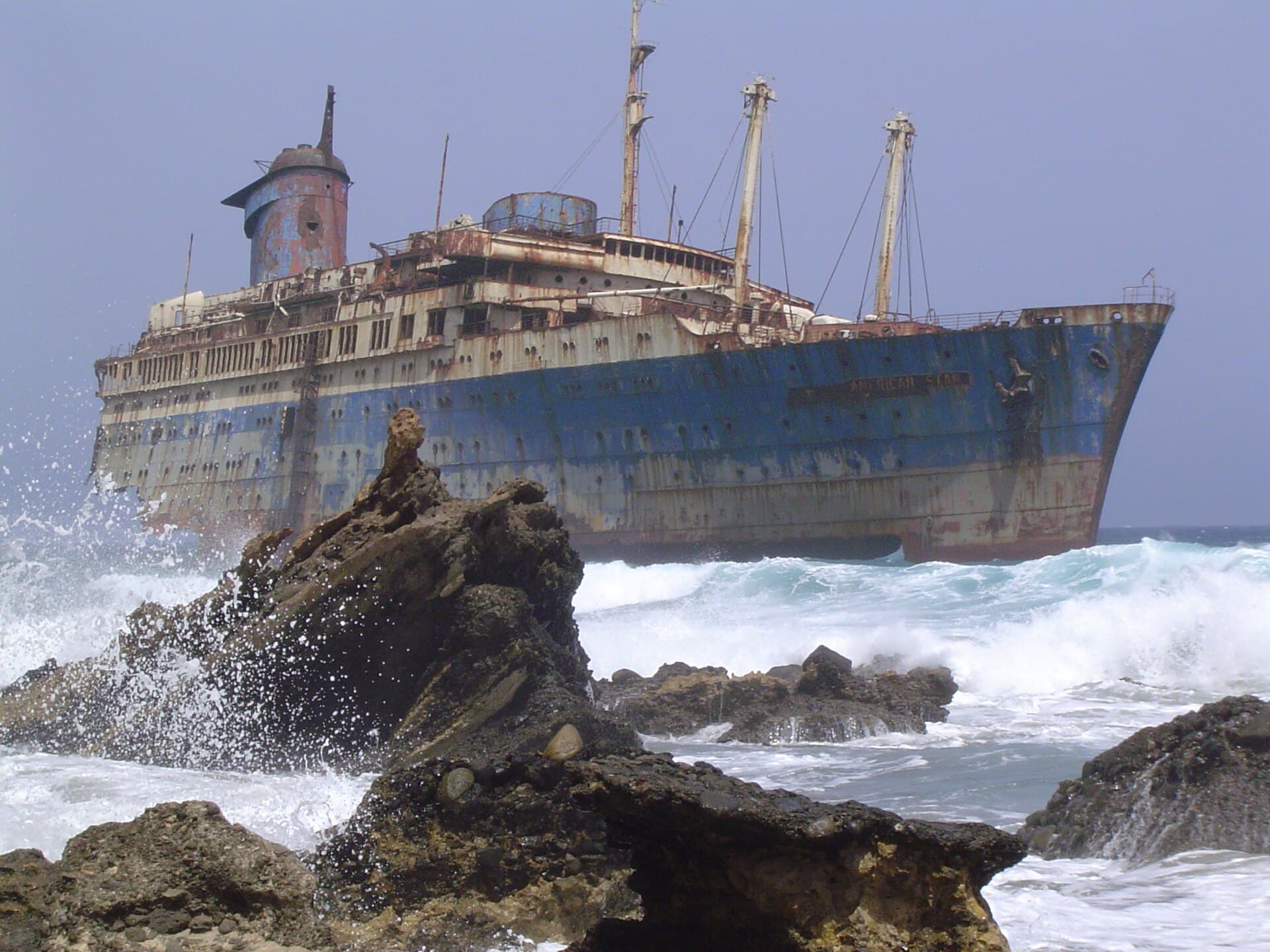 O navă mare, veche, eșuată sau abandonată lângă țărm, cu valuri care lovesc stâncile. (Foto de arhivă: Wollex / CC BY-SA 3.0, Wikimedia Commons)