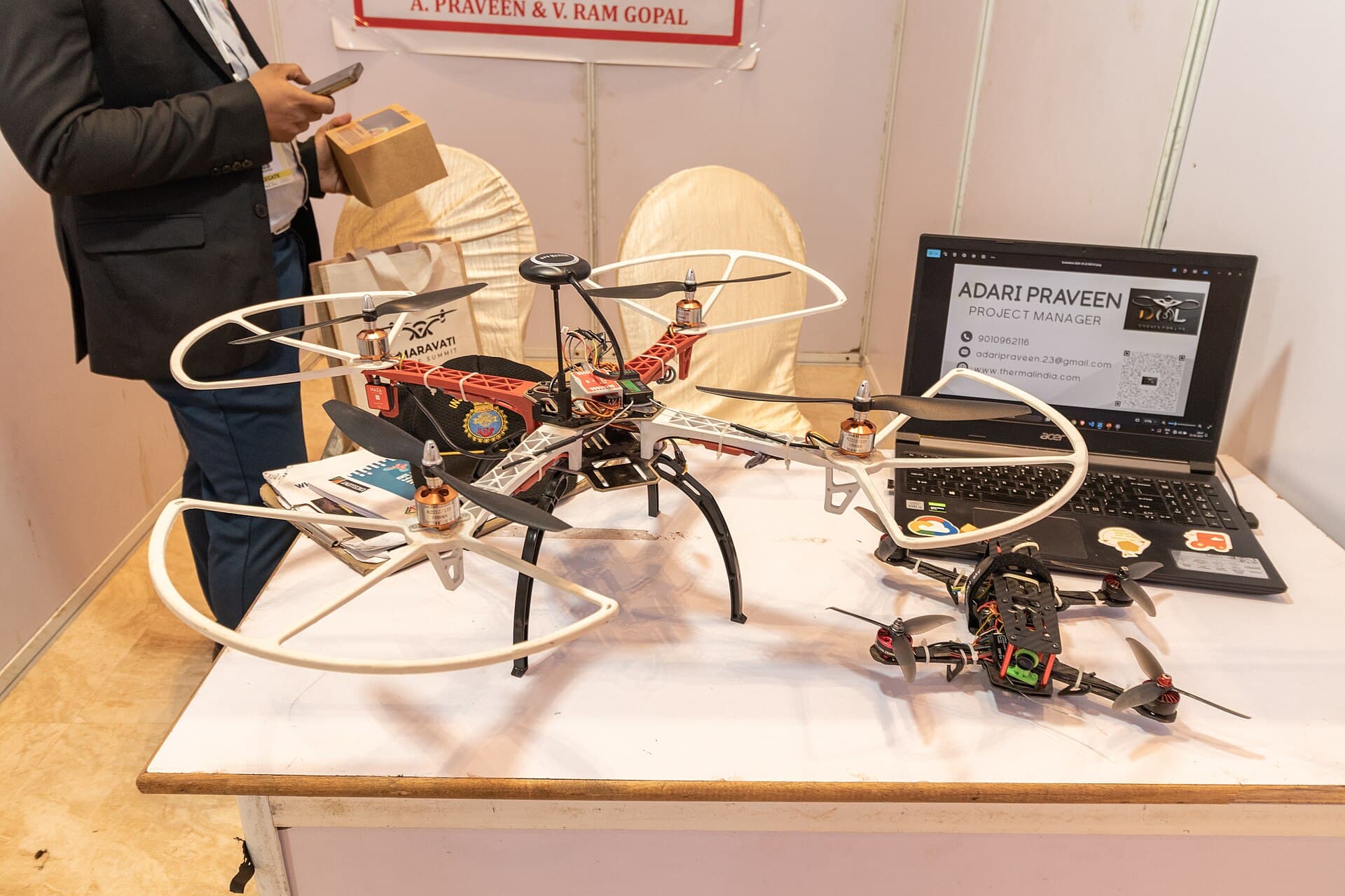 Două drone multicoptere expuse pe o masă, într-un spațiu interior, alături de un laptop și materiale de prezentare. (Foto de arhivă: iMahesh / CC BY-SA 4.0, Wikimedia Commons)
