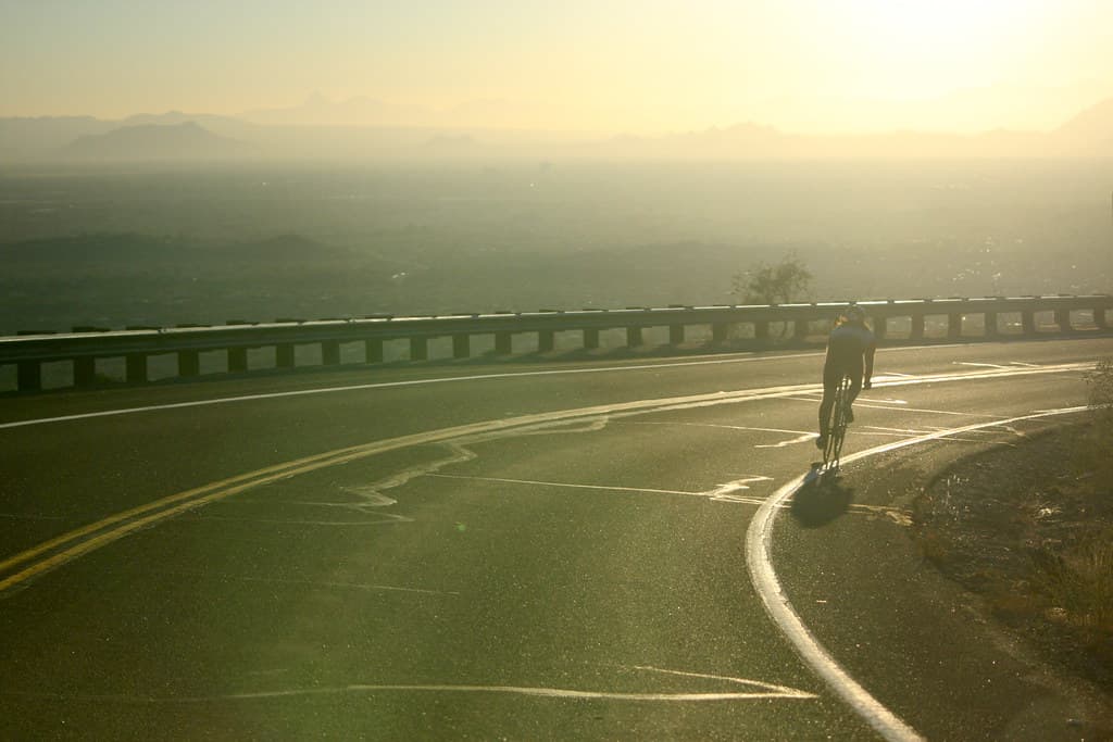 O șosea asfaltată, curbată, cu parapet, văzută într-un peisaj montan, și un biciclist. (Foto de arhivă: kevin dooley / BY 2.0, Openverse)