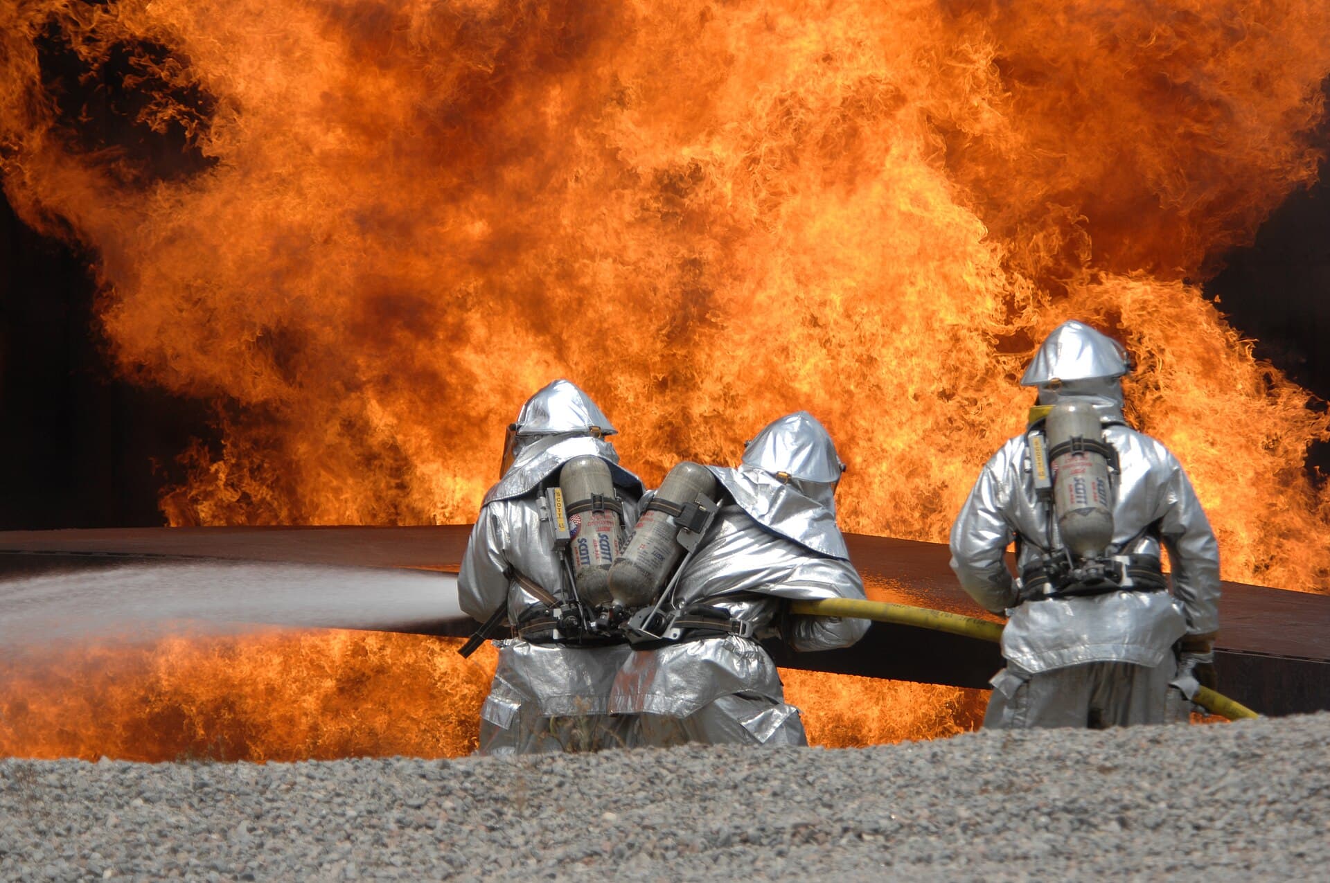 Trei pompieri în costume de protecție și cu aparate de respirat, folosind un furtun în fața unui incendiu cu flăcări mari. (Foto de arhivă: Airman 1st Class Kathrine McDowell, U.S. Air Force / Public domain, Wikimedia Commons)