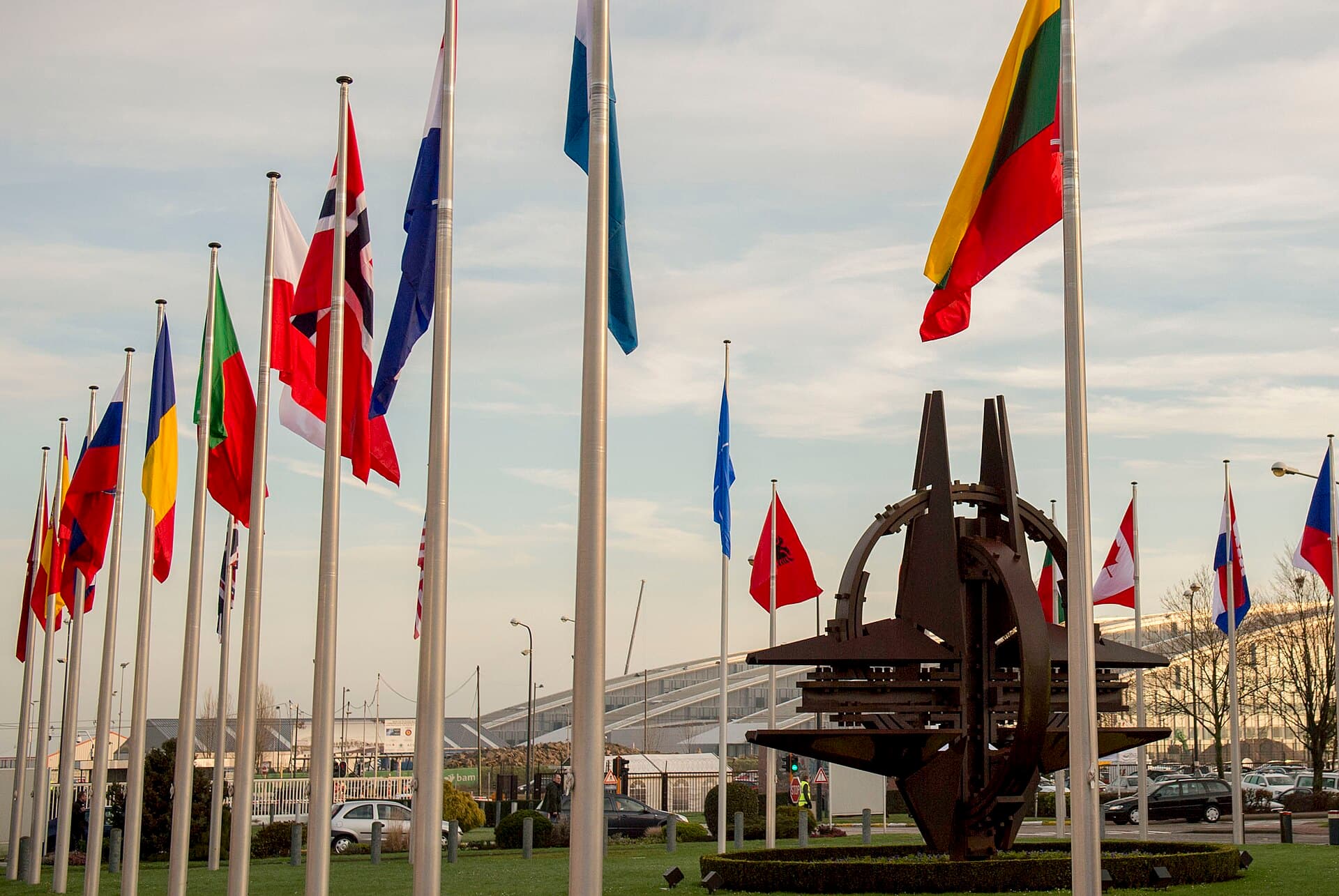 Un rând de steaguri naționale și un monument metalic într-un spațiu exterior, probabil în fața unui complex internațional. (Foto: Senior Master Sgt. Adrian Cadiz / Public domain, Wikimedia Commons)