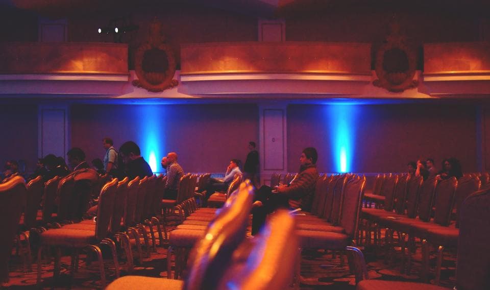 Public într-o sală de evenimente, așezat pe scaune, cu iluminare colorată pe scenă și în sală. (Foto ilustrativă: Daria Nepriakhina / CC0 1.0, Openverse)