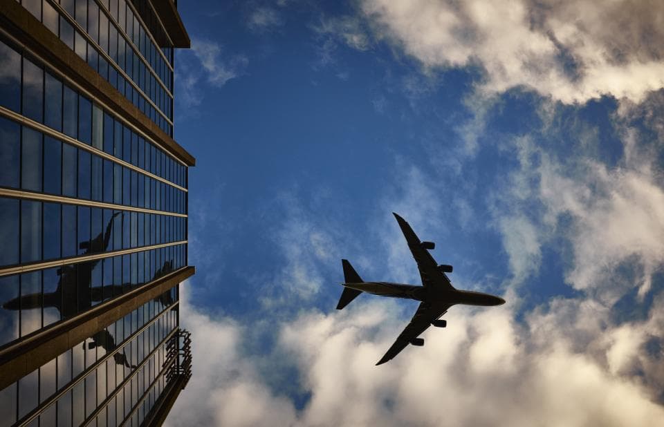 Un avion de pasageri văzut din unghi inferior, în zbor, lângă o clădire cu fațadă vitrată. (Foto ilustrativă: Robert De Bock / CC0 1.0, Openverse)