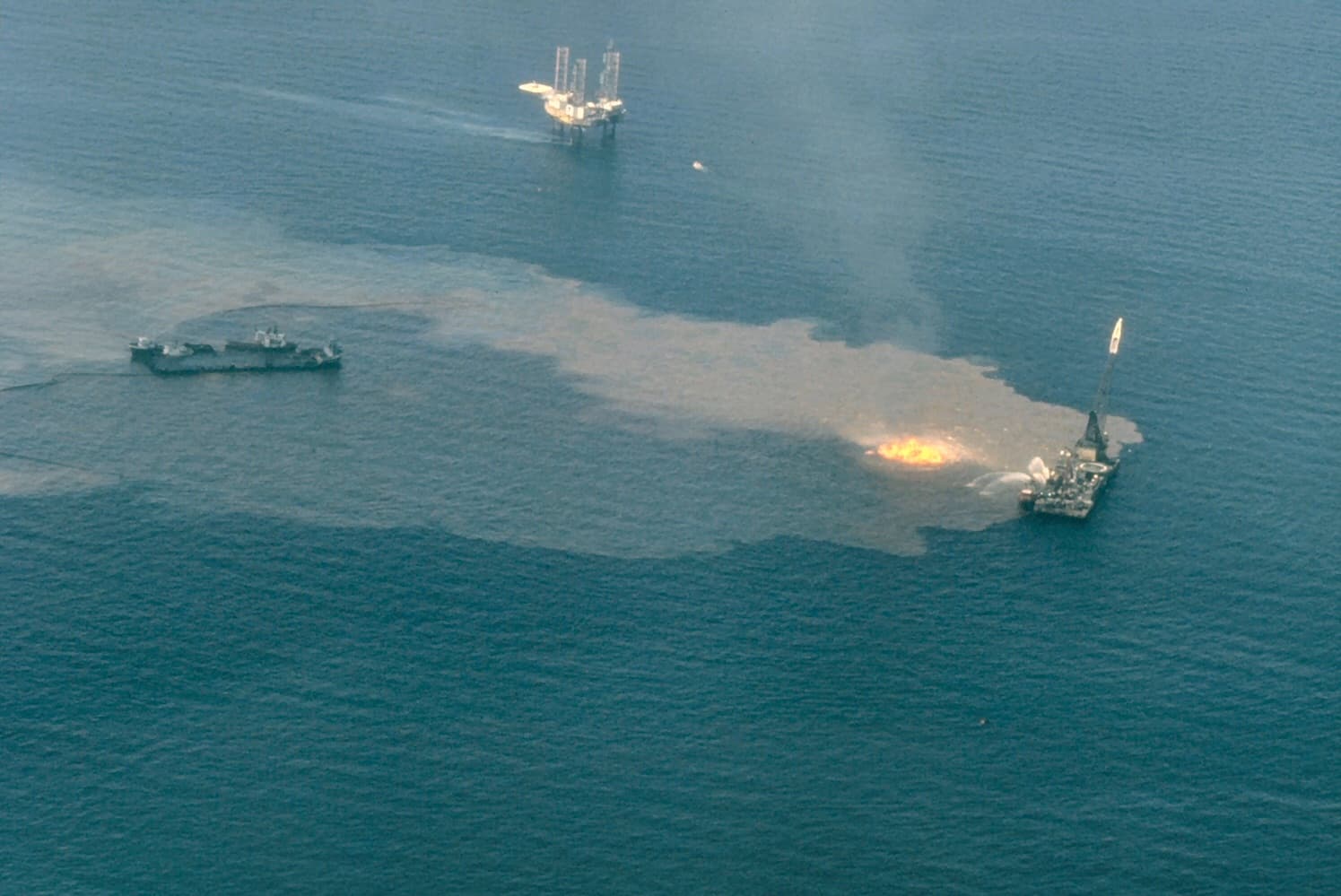O instalație sau navă industrială în larg, cu flăcări și fum la suprafața apei. (Foto de arhivă: U.S. — NOAA / Public domain, Wikimedia Commons)