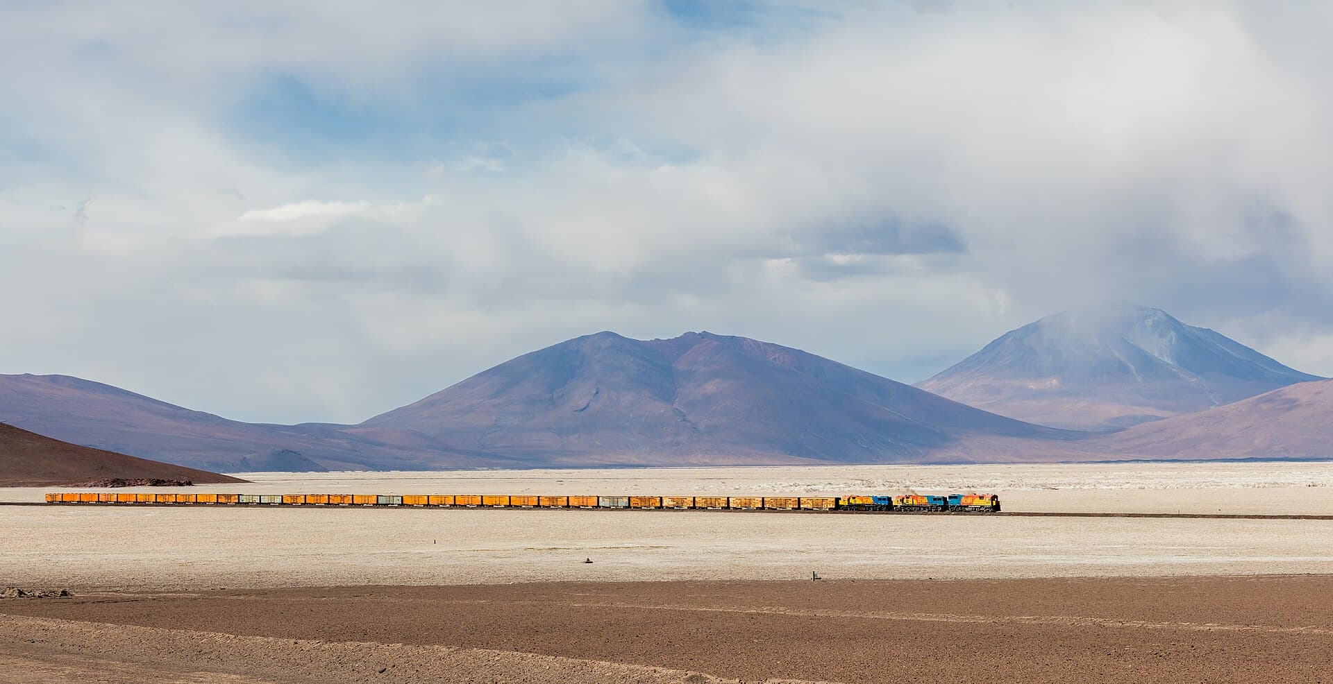 Un tren lung de marfă, cu vagoane de transport, traversând un peisaj arid cu munți. (Foto de arhivă: Diego Delso / CC BY-SA 4.0, Wikimedia Commons)