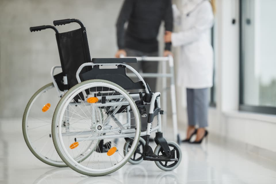 Un scaun rulant într-un spital, cu personal medical în fundal. (Foto ilustrativă: Direct Media / CC0 1.0, Openverse)