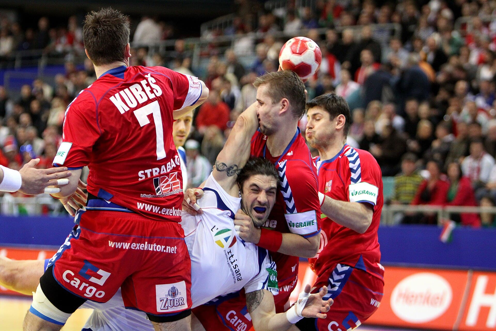 Un meci de handbal masculin, cu jucători în echipamente roșii și albe, aflați într-un duel pe teren. (Foto de arhivă: Steindy ( talk ) 00:03, 21 January 2010 (UTC) / CC BY-SA 3.0, Wikimedia Commons)