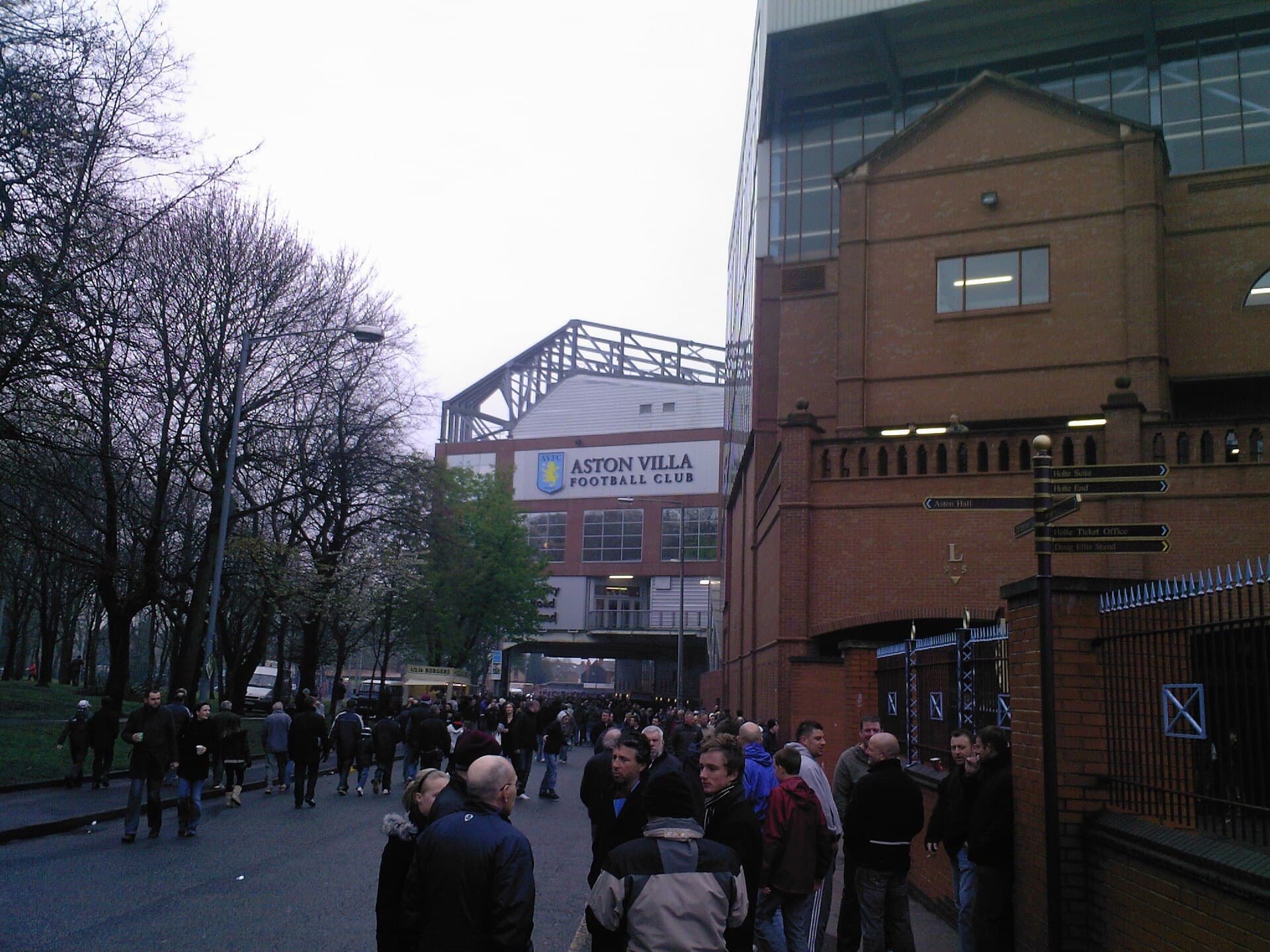 Exteriorul stadionului Aston Villa, cu mulțimi de suporteri în jurul arenei și un panou pe care scrie „Aston Villa Football Club”. (Foto: User:Woodym555 / CC BY-SA 3.0, Wikimedia Commons)